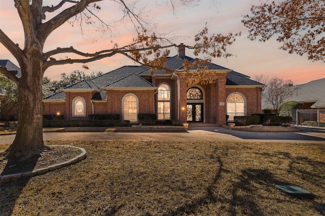 view of front of house with brick siding, french doors, a lawn, curved driveway, and a chimney