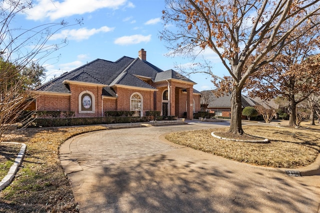 view of front facade with roof with shingles, brick siding, curved driveway, and a chimney