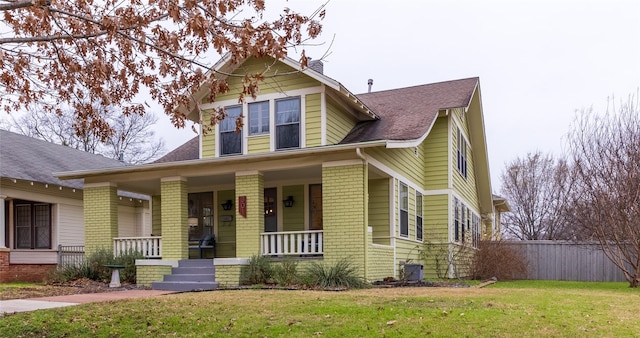 view of front facade featuring brick siding, roof with shingles, covered porch, a front yard, and cooling unit