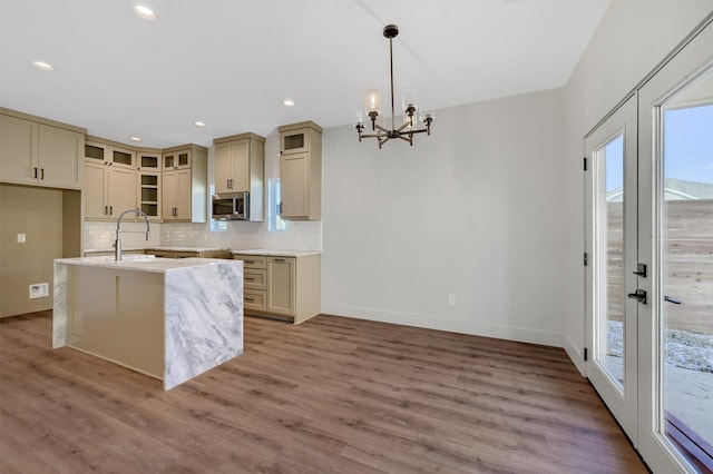 kitchen featuring a kitchen island with sink, light countertops, stainless steel microwave, glass insert cabinets, and pendant lighting