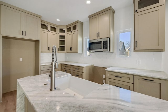 kitchen featuring stainless steel microwave, backsplash, glass insert cabinets, and light stone countertops