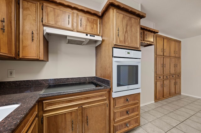 kitchen with white oven, brown cabinetry, light tile patterned floors, under cabinet range hood, and black electric cooktop