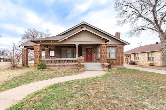 bungalow with covered porch, brick siding, a chimney, and a front lawn