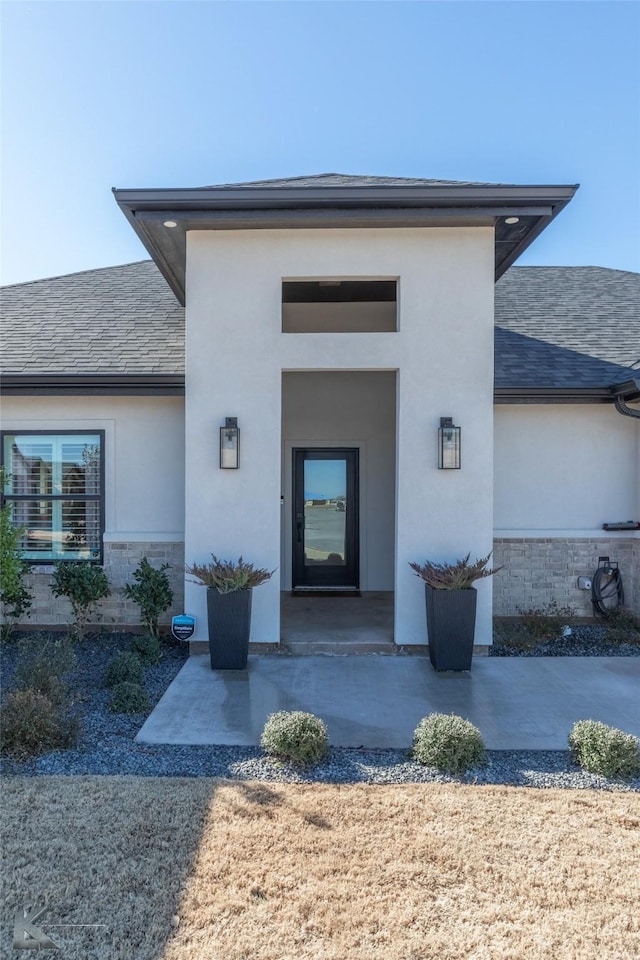 entrance to property featuring stone siding, roof with shingles, a patio area, and stucco siding