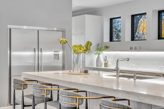 kitchen with a sink, white cabinets, light stone countertops, stainless steel built in refrigerator, and modern cabinets