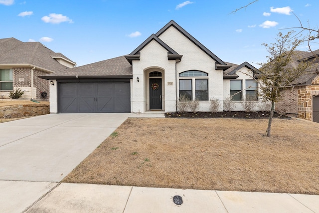 french country inspired facade with concrete driveway, brick siding, an attached garage, and roof with shingles