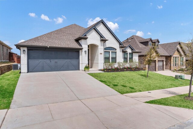 french country inspired facade with roof with shingles, central AC unit, an attached garage, and brick siding
