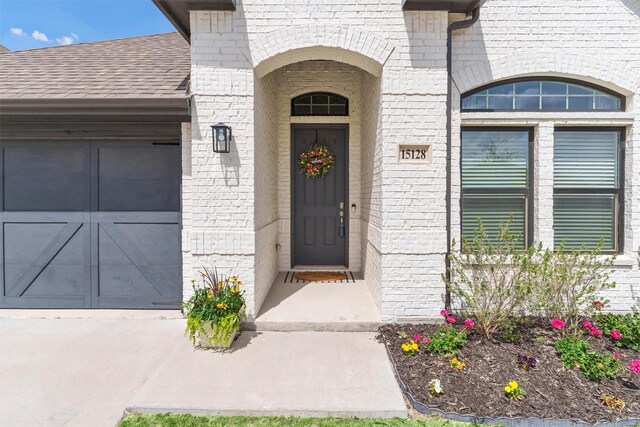 view of exterior entry featuring roof with shingles, an attached garage, concrete driveway, and brick siding