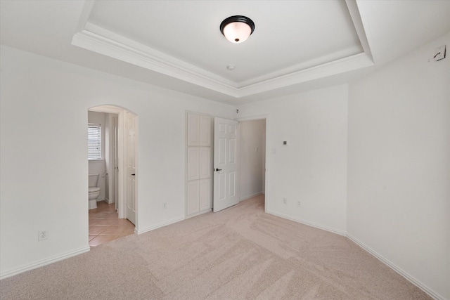 unfurnished bedroom with a tray ceiling, arched walkways, light carpet, ensuite bath, and baseboards