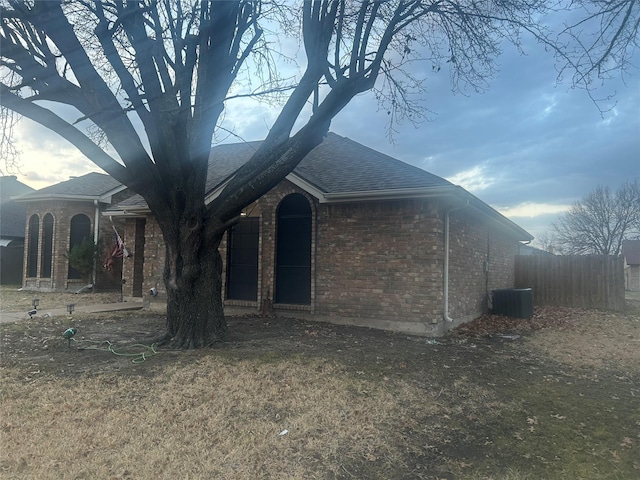 view of front facade featuring brick siding, roof with shingles, fence, and central AC unit