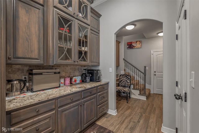 kitchen with arched walkways, dark brown cabinetry, light stone countertops, dark wood finished floors, and glass insert cabinets