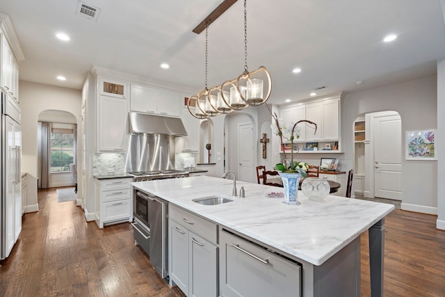 kitchen featuring arched walkways, under cabinet range hood, visible vents, white cabinets, and an island with sink