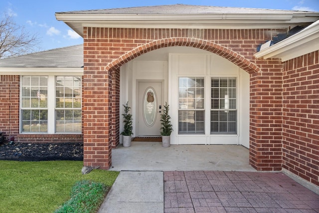 property entrance featuring a patio area, roof with shingles, and brick siding