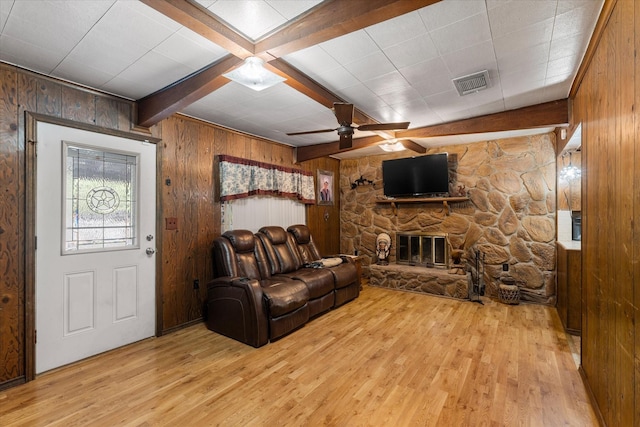 living room with a ceiling fan, a stone fireplace, light wood finished floors, wood walls, and beamed ceiling