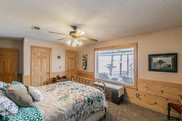 carpeted bedroom featuring a textured ceiling, ceiling fan, wood walls, visible vents, and wainscoting