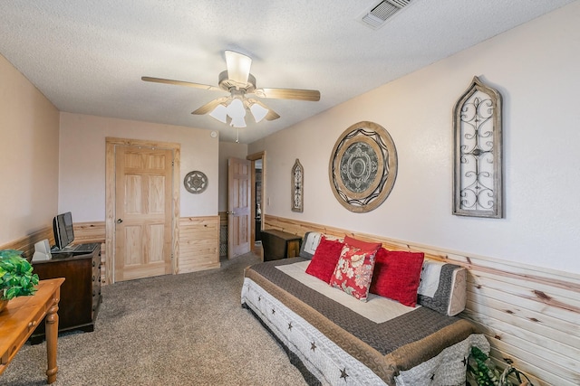 carpeted bedroom with a textured ceiling, ceiling fan, wooden walls, visible vents, and wainscoting