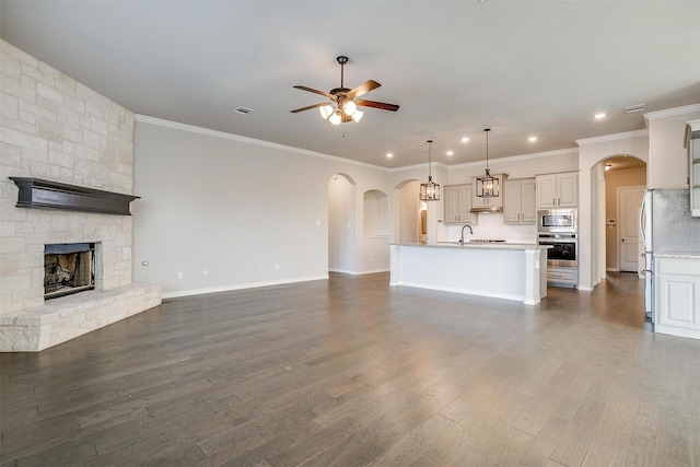 unfurnished living room with arched walkways, ceiling fan, a stone fireplace, a sink, and dark wood finished floors