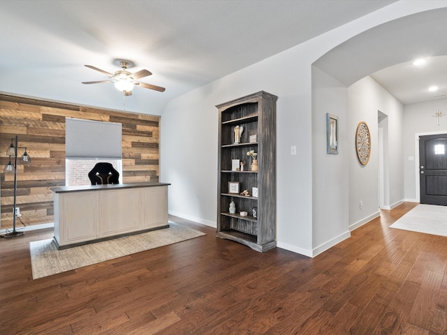 unfurnished office with arched walkways, dark wood-style flooring, wood walls, a ceiling fan, and baseboards