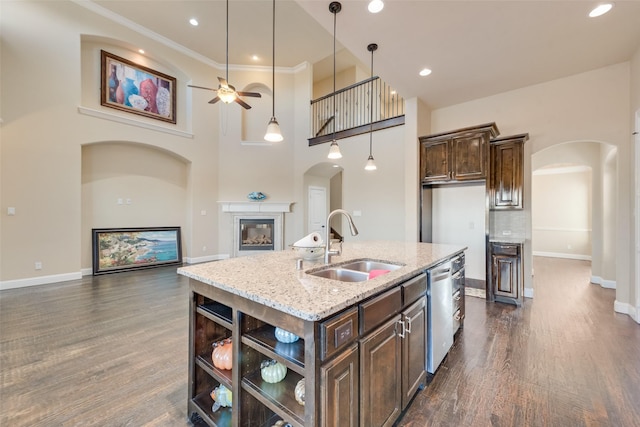 kitchen featuring arched walkways, a kitchen island with sink, a sink, dark brown cabinets, and light stone countertops