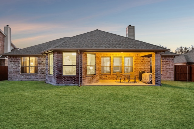 back of house at dusk featuring roof with shingles, brick siding, a chimney, a lawn, and fence