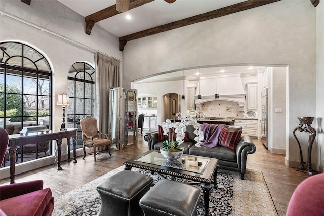 living room with arched walkways, a towering ceiling, a ceiling fan, light wood-style floors, and beam ceiling