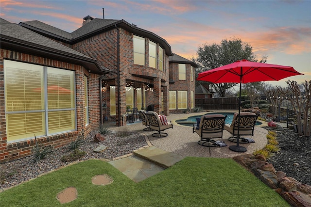 back of property at dusk with brick siding, fence, roof with shingles, a fenced in pool, and a patio area