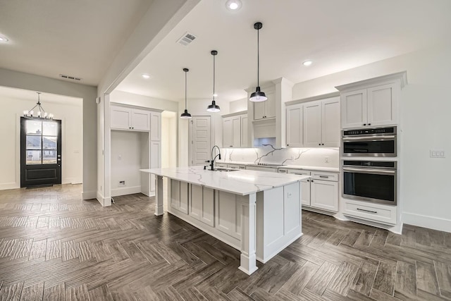 kitchen with light stone counters, stainless steel double oven, a kitchen island with sink, and decorative light fixtures