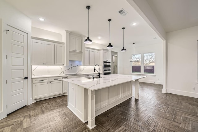 kitchen featuring a kitchen island with sink, light stone countertops, visible vents, white cabinetry, and decorative backsplash