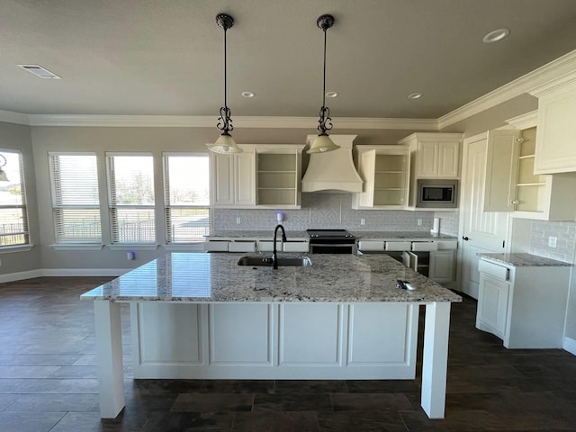 kitchen featuring a center island with sink, stainless steel microwave, pendant lighting, open shelves, and a sink