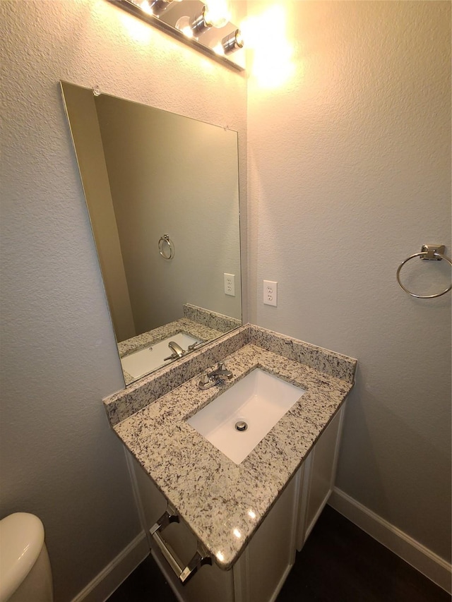 half bath featuring toilet, a textured wall, vanity, and baseboards