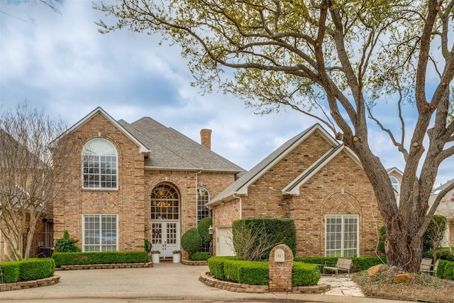 traditional-style home featuring an attached garage, a shingled roof, a chimney, french doors, and brick siding