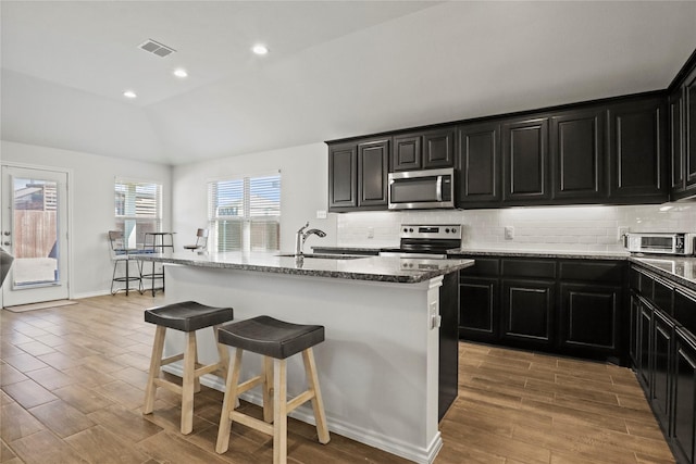 kitchen with stone countertops, a kitchen island with sink, stainless steel appliances, a sink, and a kitchen breakfast bar