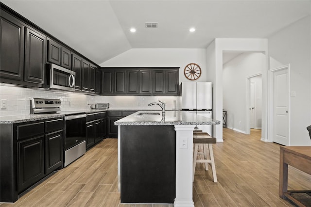 kitchen featuring visible vents, appliances with stainless steel finishes, an island with sink, dark cabinetry, and a kitchen breakfast bar