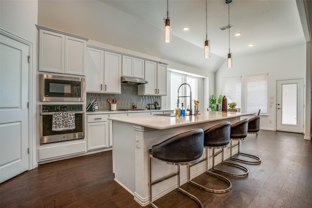 kitchen with a kitchen island with sink, under cabinet range hood, oven, light countertops, and pendant lighting