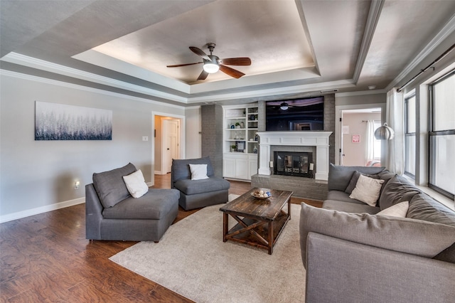 living room with dark wood finished floors, a raised ceiling, ornamental molding, a brick fireplace, and baseboards