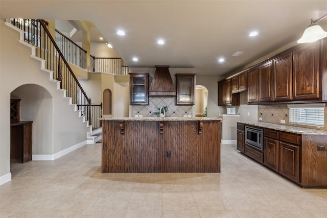 kitchen featuring arched walkways, custom exhaust hood, a breakfast bar area, stainless steel microwave, and glass insert cabinets