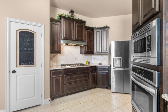 kitchen featuring light stone counters, stainless steel appliances, tasteful backsplash, dark brown cabinetry, and under cabinet range hood