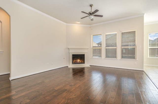 unfurnished living room featuring a warm lit fireplace, dark wood-type flooring, a ceiling fan, baseboards, and crown molding