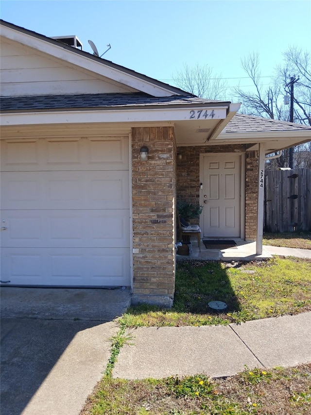 doorway to property featuring a garage, brick siding, roof with shingles, and fence