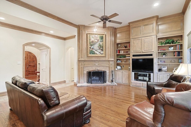 living area featuring arched walkways, ceiling fan, crown molding, light wood-type flooring, and a fireplace