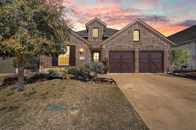 french country style house with an attached garage, brick siding, a yard, concrete driveway, and roof with shingles