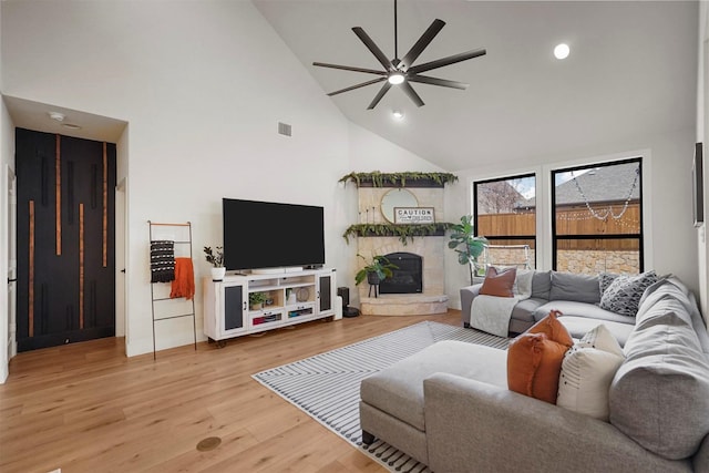 living area featuring high vaulted ceiling, a stone fireplace, visible vents, a ceiling fan, and light wood-type flooring