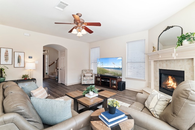 living room with arched walkways, a tile fireplace, ceiling fan, stairway, and wood finished floors