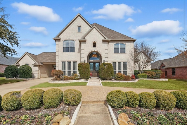 french country inspired facade featuring brick siding, driveway, french doors, roof with shingles, and a front yard