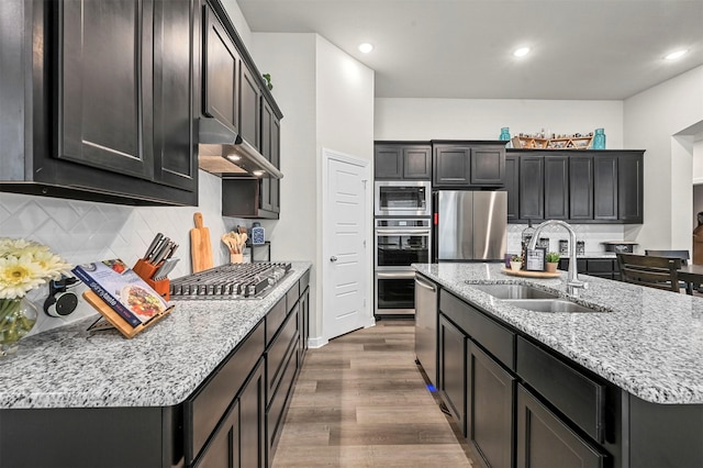 kitchen with an island with sink, dark wood-type flooring, stainless steel appliances, under cabinet range hood, and a sink