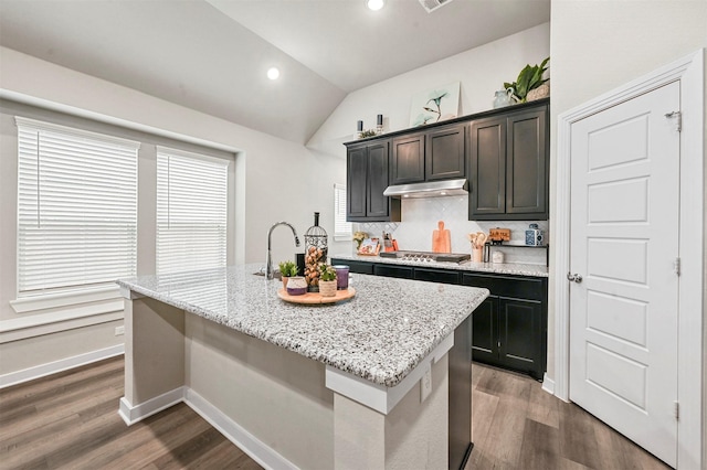 kitchen with dark wood-style floors, light stone countertops, a kitchen island with sink, vaulted ceiling, and under cabinet range hood