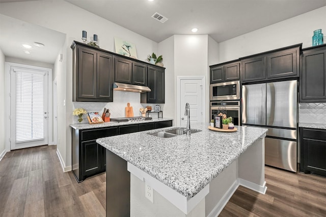 kitchen featuring light stone counters, a center island with sink, appliances with stainless steel finishes, a sink, and under cabinet range hood