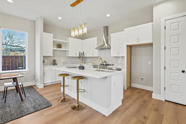 kitchen featuring white cabinets, an island with sink, wall chimney exhaust hood, decorative light fixtures, and light countertops