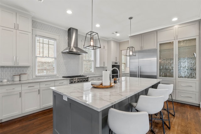 kitchen featuring white cabinets, wall chimney range hood, appliances with stainless steel finishes, a center island with sink, and pendant lighting