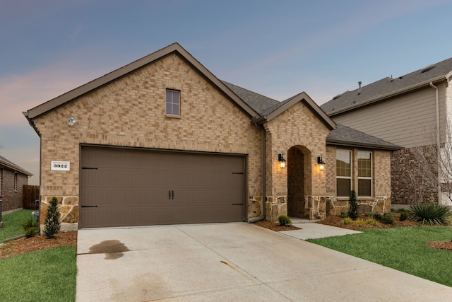 french provincial home featuring brick siding, a shingled roof, concrete driveway, an attached garage, and a front yard